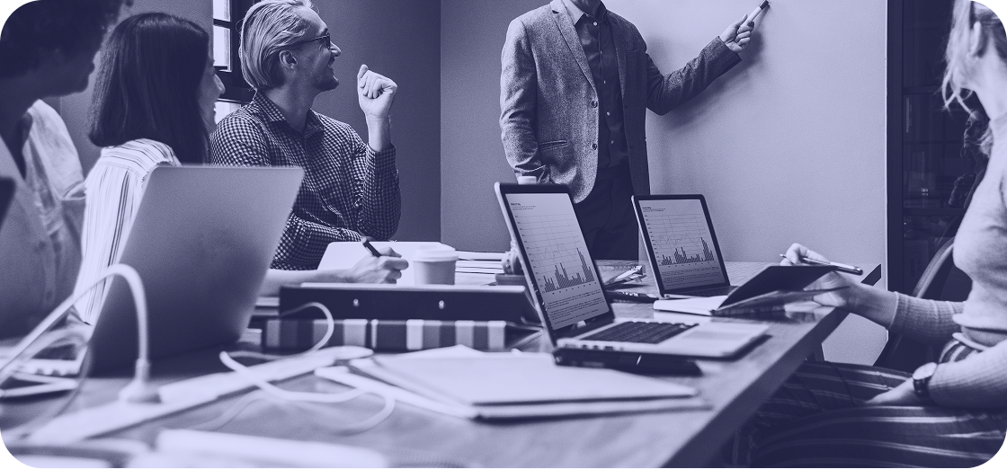 Reunión de trabajo con laptops y una persona parada explicando en una pizarra 